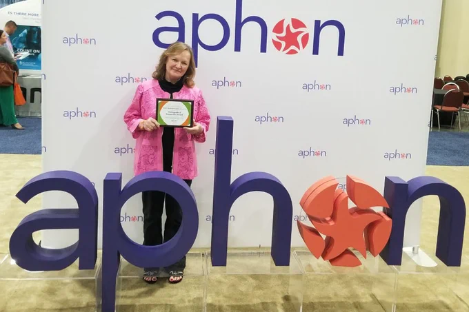 Verna Hendricks-Ferguson, Ph.D., stands in front of a sign reading "aphon" as she holds an award.