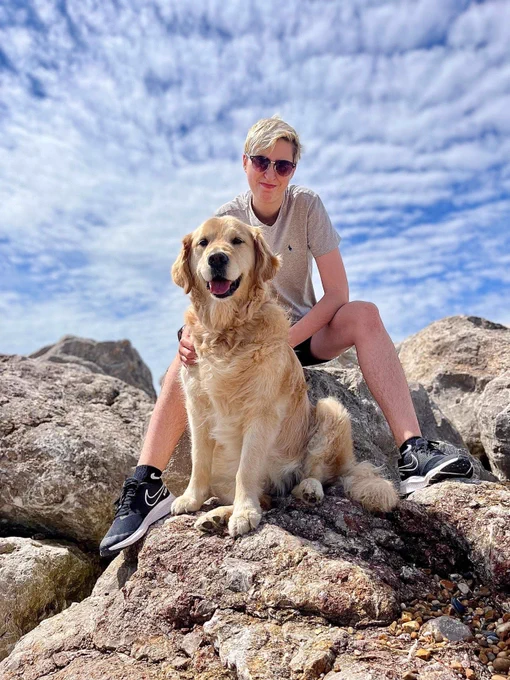 A photograph of Riley posed behind Yashka, his Guide Dog. They are sitting on rocks at Felixstowe Beach on a sunny day.