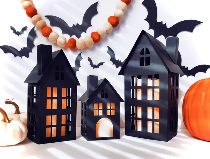 three lighted black paper houses displayed with bats, white and orange pumpkins, and felt ball garland