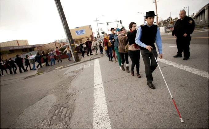 A man with a red and white stick leads a long procession of at least twenty people across a road. The man is out front and in the middle of the road. The line is long and files along the pavement, before turning at 90 degrees onto the road. Each person the line holds on to the person in front by placing their arm on their shoulder. A man in uniform unconnected by touch to the others, likely a security guard, looks on as the line of people crosses the road.