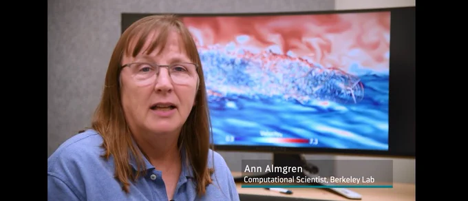 In the forefront, Ann Almgren, with light brown hair and glasses, is wearing a blue button-up shirt. In the background, a simulation of a coastal wind turbine shows on a computer monitor. 
