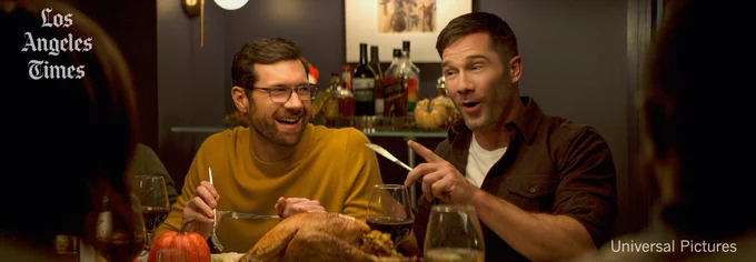 This photo from the movie "Bros" shows two men animatedly sitting around a dinner table at a dinner party. There is a bar cart with bottles in the background.