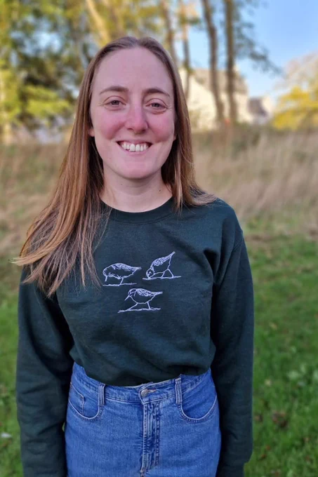 Blond woman in a forest green sweater with three embroidered sanderlings 