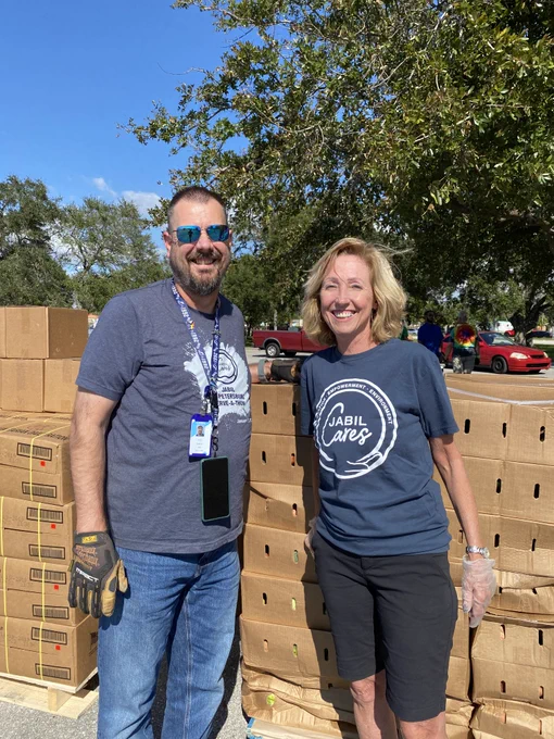 A man wearing sunglasses, a navy blue T-shirt and jeans and a woman wearing a navy blue T-shirt that says "Jabil Cares" and black shorts stand in front of pallets of cardboard boxes outside in front of a tree.