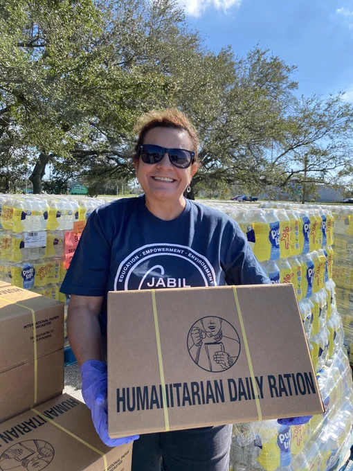 A woman with brown hair wearing sunglasses and a navy blue shirt that says "Jabil Cares" is holding a cardboard box that reads "Humanitarian Daily Ration" in front of a pallet of water bottles.