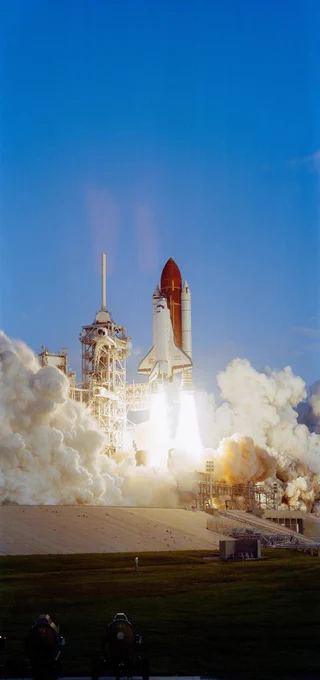 A space shuttle, attached to a rocket, launching to space and detaching from the launch pad. Smoke fills the the surrounding area.