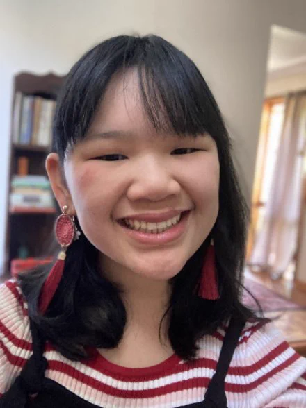  Selfie of Paige, a young Chinese Australian person, smiling at the camera. They have a fringe and dark hair just past their shoulders. They are wearing dangly red earrings and a red and white striped top.