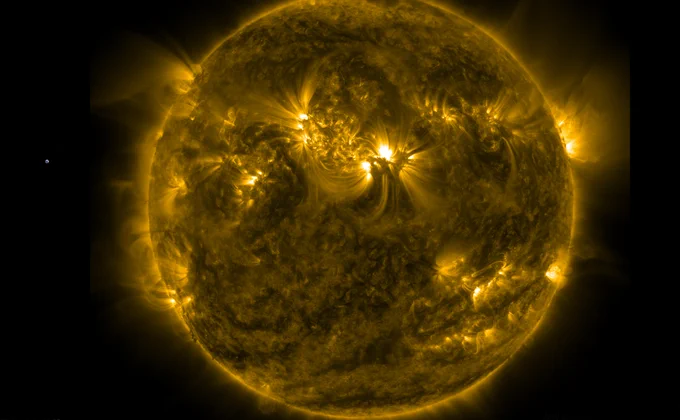 Die Sonne in gelber Darstellung. Eine Aktive Region genau in der Mitte der erdzugewandten Seite. Von diesem weißen Fleck gehen Strahlen weit in die Umgebung aus. Zum Größenvergleich links als winziger Punkt die Erde einkopiert.