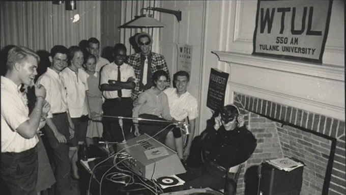 A black and white photograph from the mid-60s shows a WTUL DJ broadcasting live surrounded by a group of students.