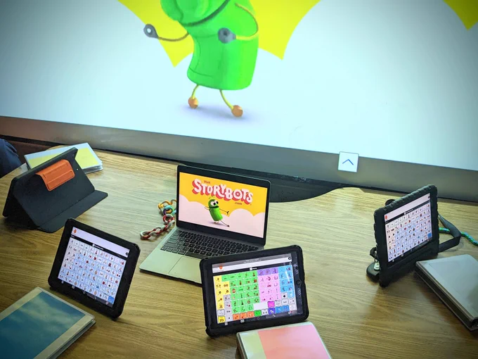 4 iPads, with Proloquo 2 Go, and 4 binders around a table with a laptop in the middle. On the laptop is Storybots, and the same image is projected on the screen behind the table.