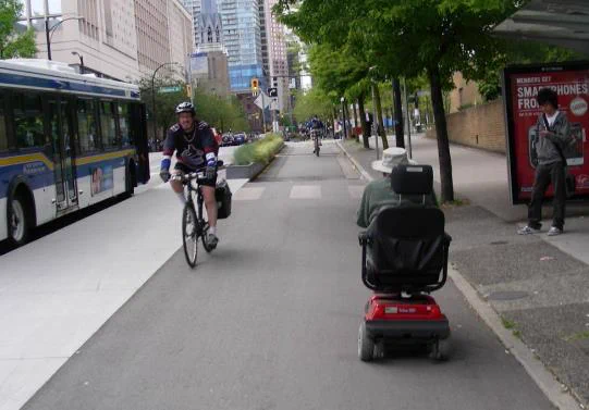 In this street scene of downtown Vancouver, a person on the right side of the image stands on the sidewalk under a bus shelter reading their phone. There is a cycle lane to the left of the bus shelter and a person is using a red and black mobility scooter to travel away from the viewer.  To their left a person using a bicycle is traveling towards the view.  A sidewalk is to the bike path's left. at the left edge of this second sidewalk, a city bus is stopped with the doors open to accept passengers.  A pedestrian walkway with white lines goes from one sidewalk to the other to facilitate pedestrian travel across the cycle lane to reach the bus from the bus shelter.