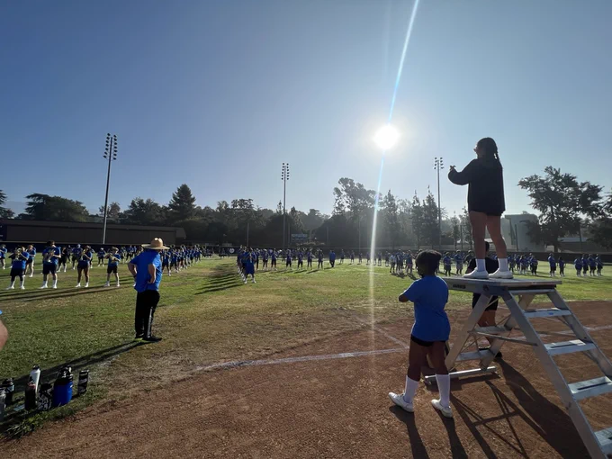 Photo of the UCLA Marching Band rehearsing on a field with the rising sun behind them. 