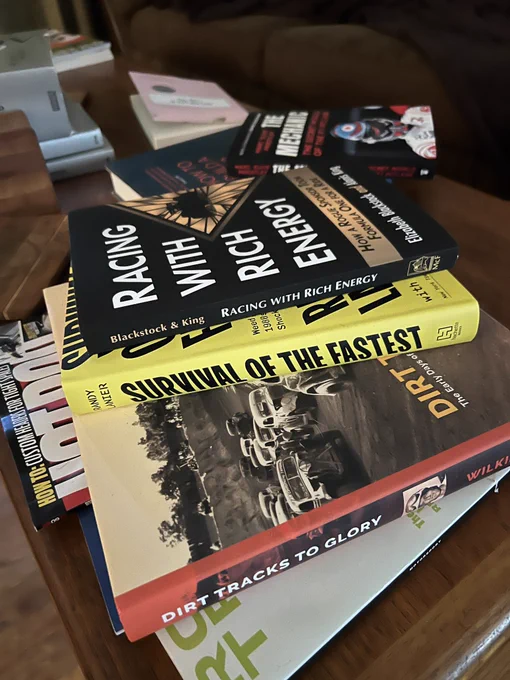 A pile of books on a wooden table including Racing with Rich Energy, Survival of the Fastest, and Dirt Tracks to Glory. Underneath the books is the Racing Issue of Road and Track Magazine