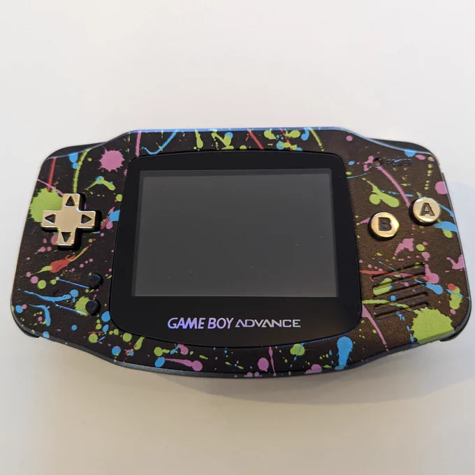 A Game Boy Advance, the shell looks like it's paint splattered in lots of colours with a black background. The buttons are brass.