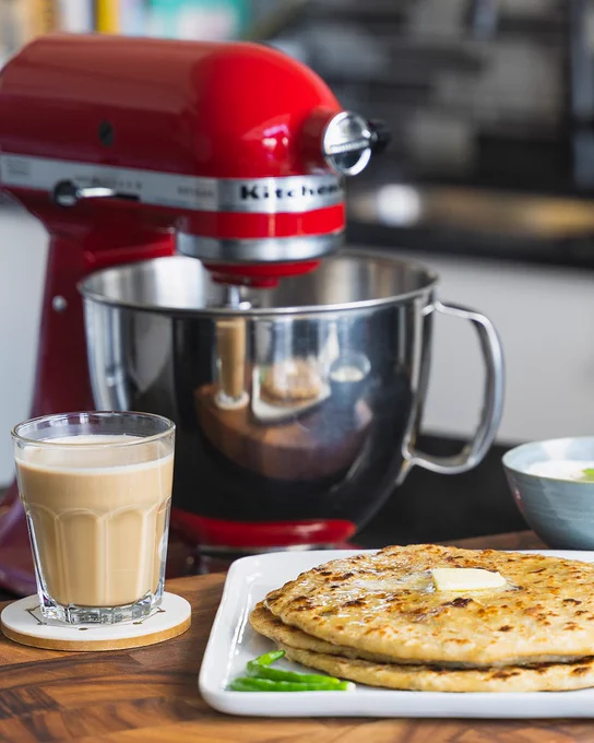 Aloo Paratha - A South Asian dish is plated on the counter with butter on top. There's a glass of milk tea on the side, and the red KitchenAid Stand Mixer with Bowl sits on the background.