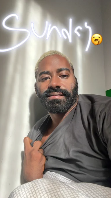 A black man looking in the camera against a stream of light. His left hand clutching his shirt