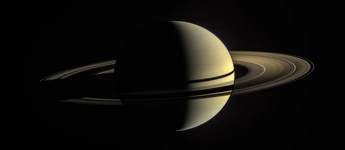 A view of Saturn taken by the Cassini spacecraft. Its right side is lit up by the Sun and the left side is a shadow, giving the planet a crescent shape. Its rings are also visible.