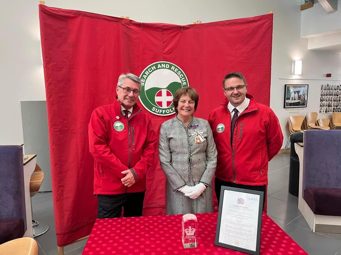 Three people standing in front of a red backdrop, which reads "Search and Rescue Suffolk", and behind a table on which stands an award and certificate.