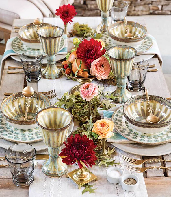 A tablescape of soft blue, sage, and cream earthenware punctuated with matching goblets and red, pink, and peach flowers, arranged on a bleached wooden table.