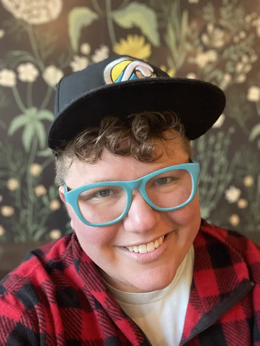 A selfie of a cute white person wearing a buffalo plaid jacket, chunky teal framed glasses, and a black snapback over their short curly dark blonde hair.