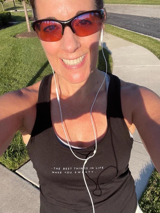 Deb Cupp, a woman with brown hair and brown eyes, takes a selfie of herself during a run. She is smiling and wearing a tank top that says, “The best things in life make you sweaty”