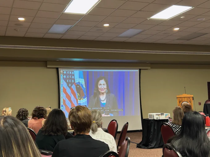 Secretary of the Interior, Deb Haaland speaks to SWAF 2022 attendees. Sec. Haaland is seen on the front wall of a room on a projector screen. The back of attendees' heads can be seen watching the video. 