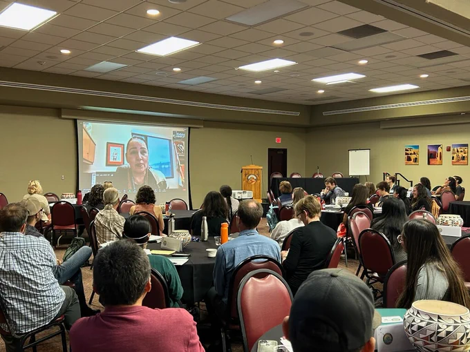 A large audience can be seen watching a woman give the Welcome Address, projected onto a screen. There is a large audience sitting at round tables. 