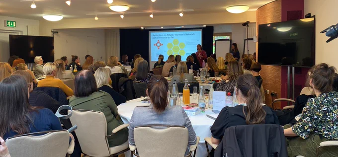 Photo shows a busy room with people sat around tables facing an interactive board.
