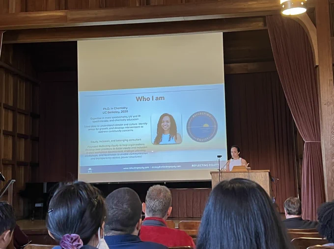 Chrissy Stachl speaking at a podium with a listening audience. There is a large screen showing a Powerpoint slide that reads 'Who I am, PhD in Chemistry, UC Berkeley 2020. Expertise in mass spectrometry, UV and IR spectroscopy, and chemistry education. Used data to understand climate and culture, identify areas for growth, and develop interventions to address community concerns. Equity, inclusion, and belonging consultant. Founded Reflecting Equity to help organizations develop best practices to foster equity and inclusion in their workplace (assessment, strategic planning, workshops, and facilitation to enable communication and transparency across power structures).'