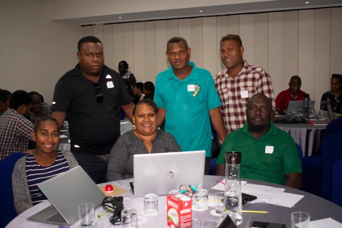 A group of people from Solomon Islands sitting around a table working at laptops. There are both men and women and they're all looking at the camera smiling. 