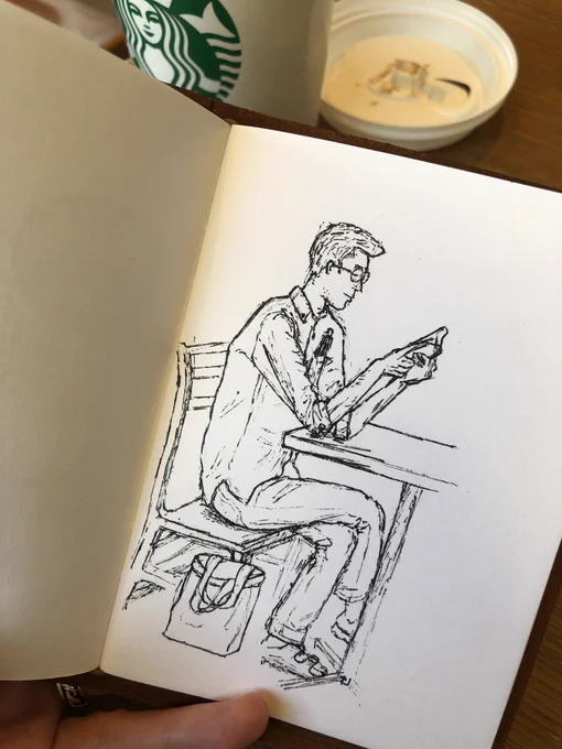 Photo of a black and white ink sketch of a young Japanese man in glasses sitting crosslegged at a table, reading a book he’s holding in his hands.