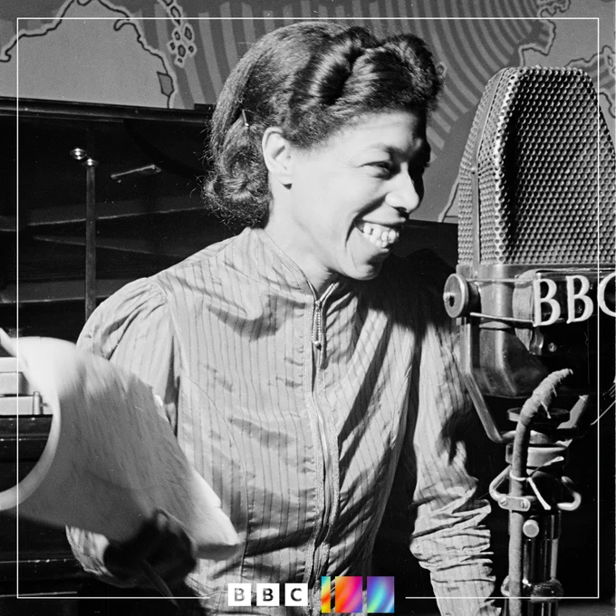 Una Marson, a Caribbean woman, is pictured in black and white standing behind a BBC microphone with a piece of paper in hand. She is laughing.