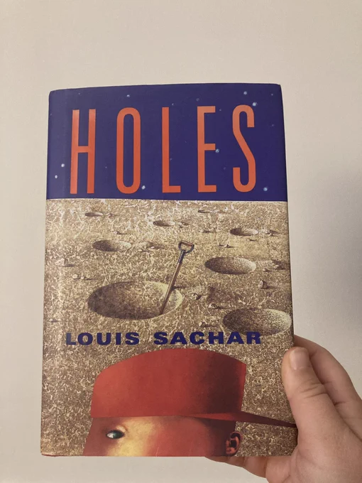 Hardcover book cover of Louis Sachar’s Holes. The side profile of a boy wearing a backwards orange cap in front of a series of perfectly circular holes dug in parched soil.