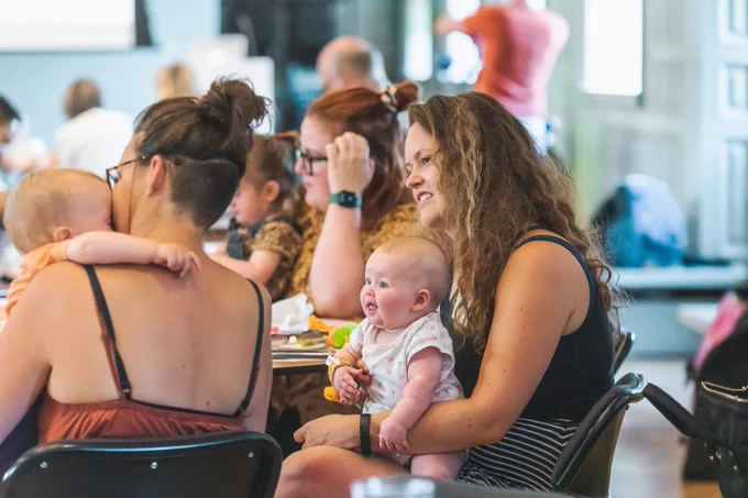 A photo of a group of mothers and their babies and children sitting at a table in a brightly lit room.