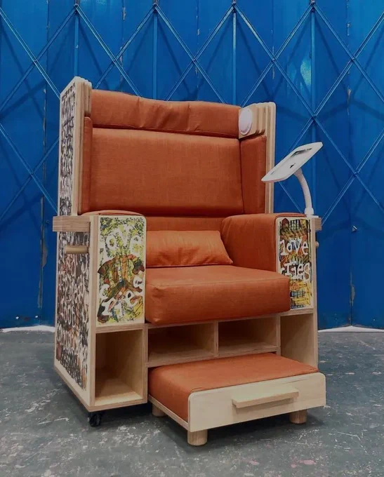 A photo of a high backed, squarish chair made from plywood and orange padding, with outer shell covered in illustrations, a screen on a stem, speakers around the head area and a pullout foodstool