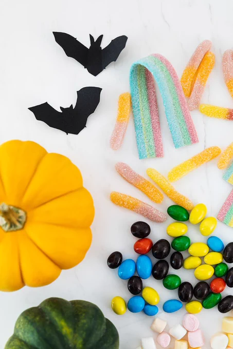 An aerial view shows an assortment of candies on a white surface; a small orange gourd and some little black bats are also visible.