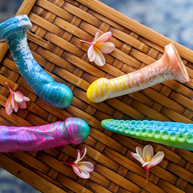 Four tropical-colored dildos, including a tentacle-shaped one, on a wicker table, arranged in a circle and shot from above.