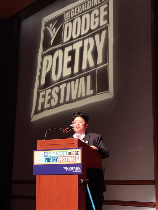 Brown round transgender writer features at Dodge poetry Festival on podium and stage. A blue cane rests against the podium, white spotlight reads above DODGE POETRY FESTIVAL