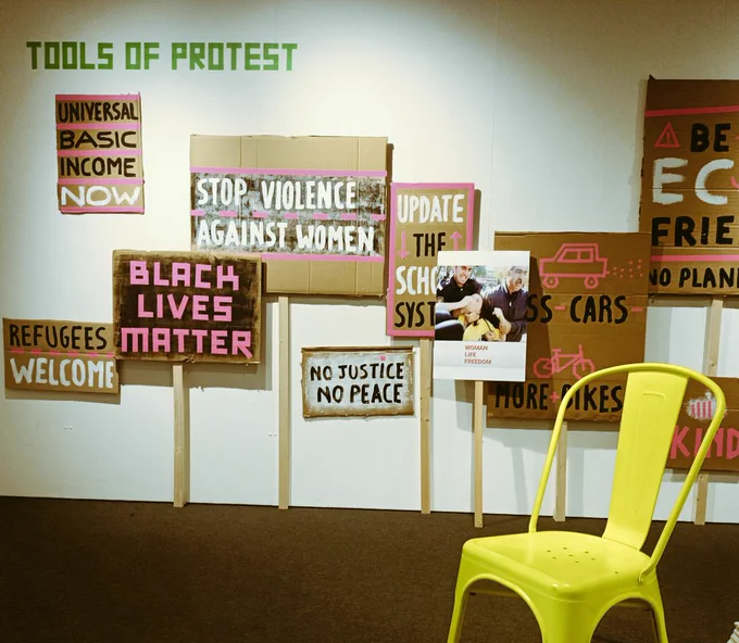 a wall of cardboard placards with pink accent words. A bright yellow chair to bottom right 