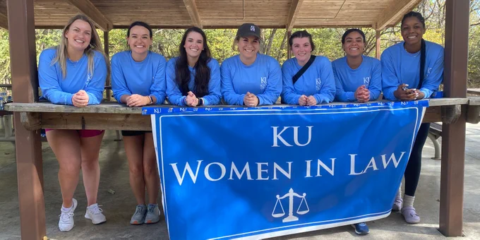 Members of Women in Law pose with a blue banner reading "KU Women in Law" at a Clinton Lake park shelter