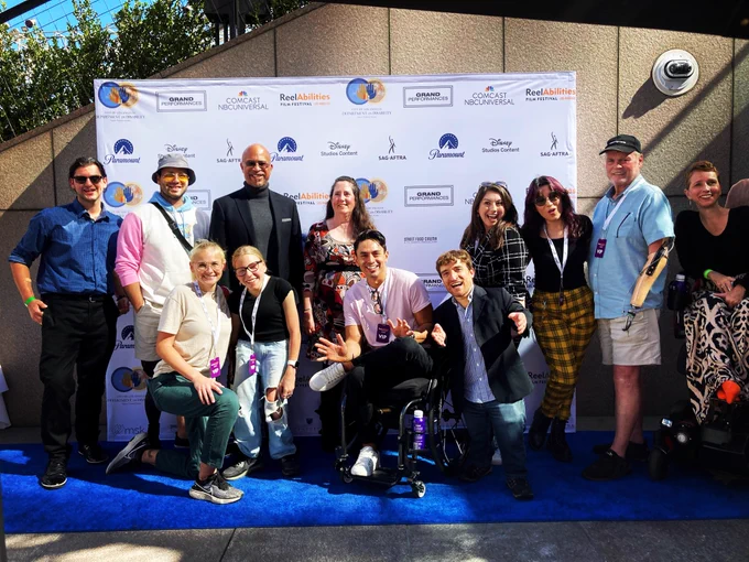 A group photo of film challenge participants on a blue carpet with a step and repeat that says reelabilities film festival Los Angeles. Everybody is smiling and it is a diverse group of film challenge participant with different disabilities.