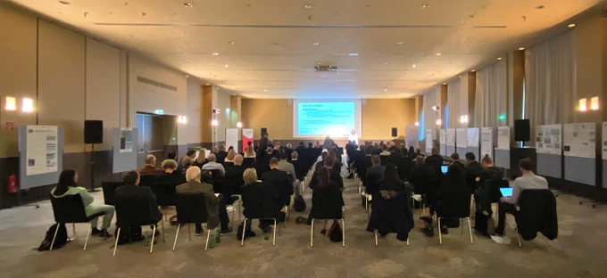 Ein Konferenzraum mit vielen Menschen auf einzelnen Sesseln, ganz hinten steht eine Person, die von starkem Licht angeleuchtet wird und hinter ihr ist eine Leinwand auf der die Worte "das Konsortium" zu lesen sind.