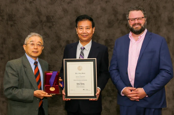 Three individuals in business attire, the one in the middle holding a plaque and one on the left holding a medal. 