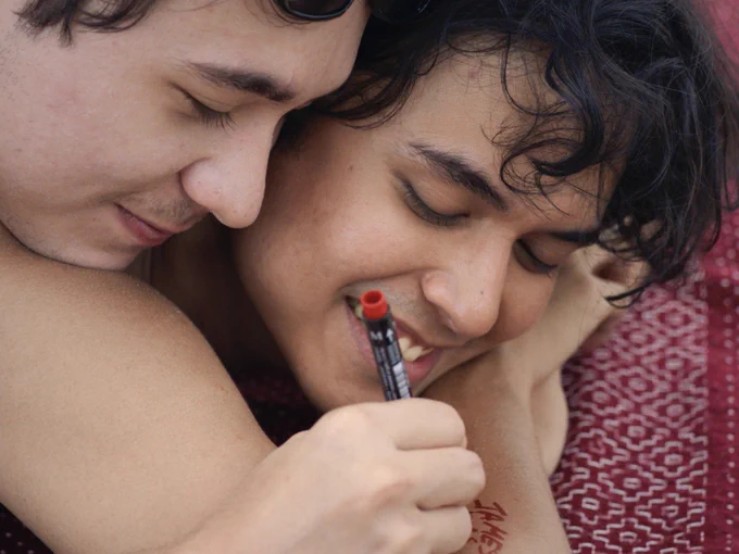 Two people are cuddling intimately. With tender joy on their face, one is writing on the other’s arm with a red marker.