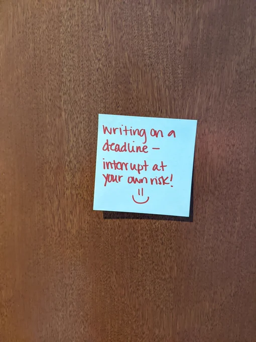 A yellow post-it note on a wooden door. In red ink, it says "writing on a deadline -- interrupt at your own risk!" It ends with a hand drawn smiley face.