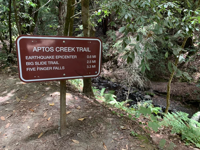 A trail sign that says "Aptos Creek Trail: Earthquake Epicenter 0.6 miles, Big Slide Trail 2.5 miles, and Five Finger Falls 3.3 miles"