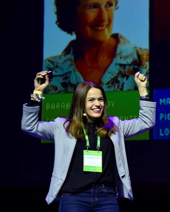 Lara giving a speech on stage, smiling with her hands in the hair wearing a black shirt and blue coat.