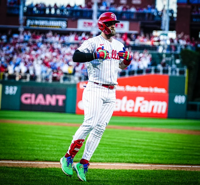Bryce Harper pointing to the Phillies logo on his chest after hitting a two-run home run. He is a Philadelphia Phillie. He's wearing the red pinstripes uniform.