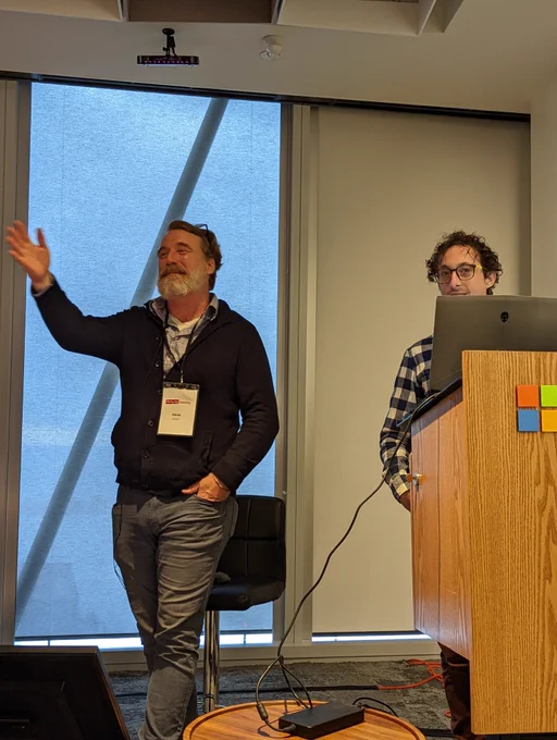Dave Evans waves from beside Microsoft branded lectern, tech standing behind.