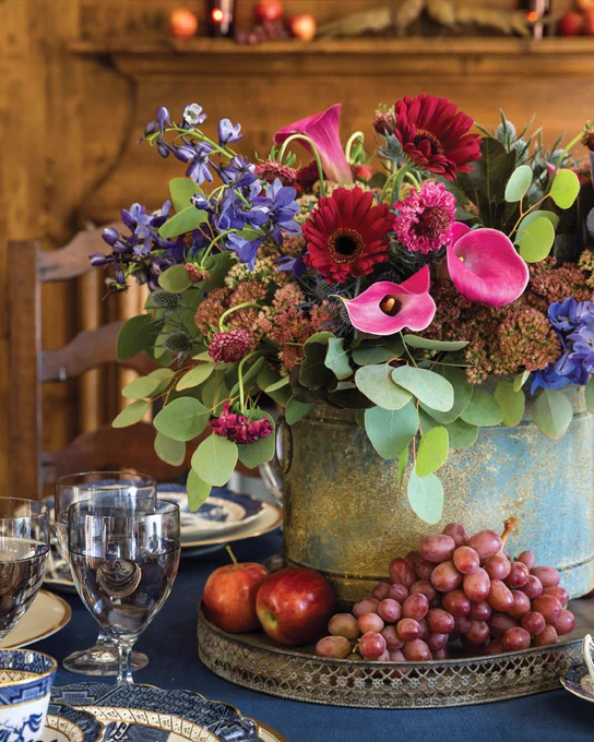 A floral arrangement of rich jewel tones sits in a metal bucket on a metal tray filled with apples and grapes.